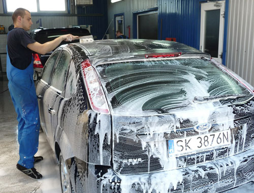 Worker applying Rain-X protective coating on a car covered in foam, enhancing shine and repelling water, dirt, and grime