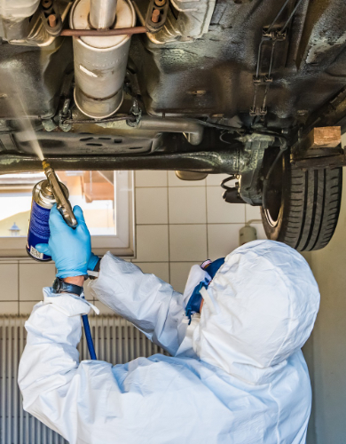 Technician in protective suit cleaning the underside of a vehicle with specialized spray equipment during underbody wash.
