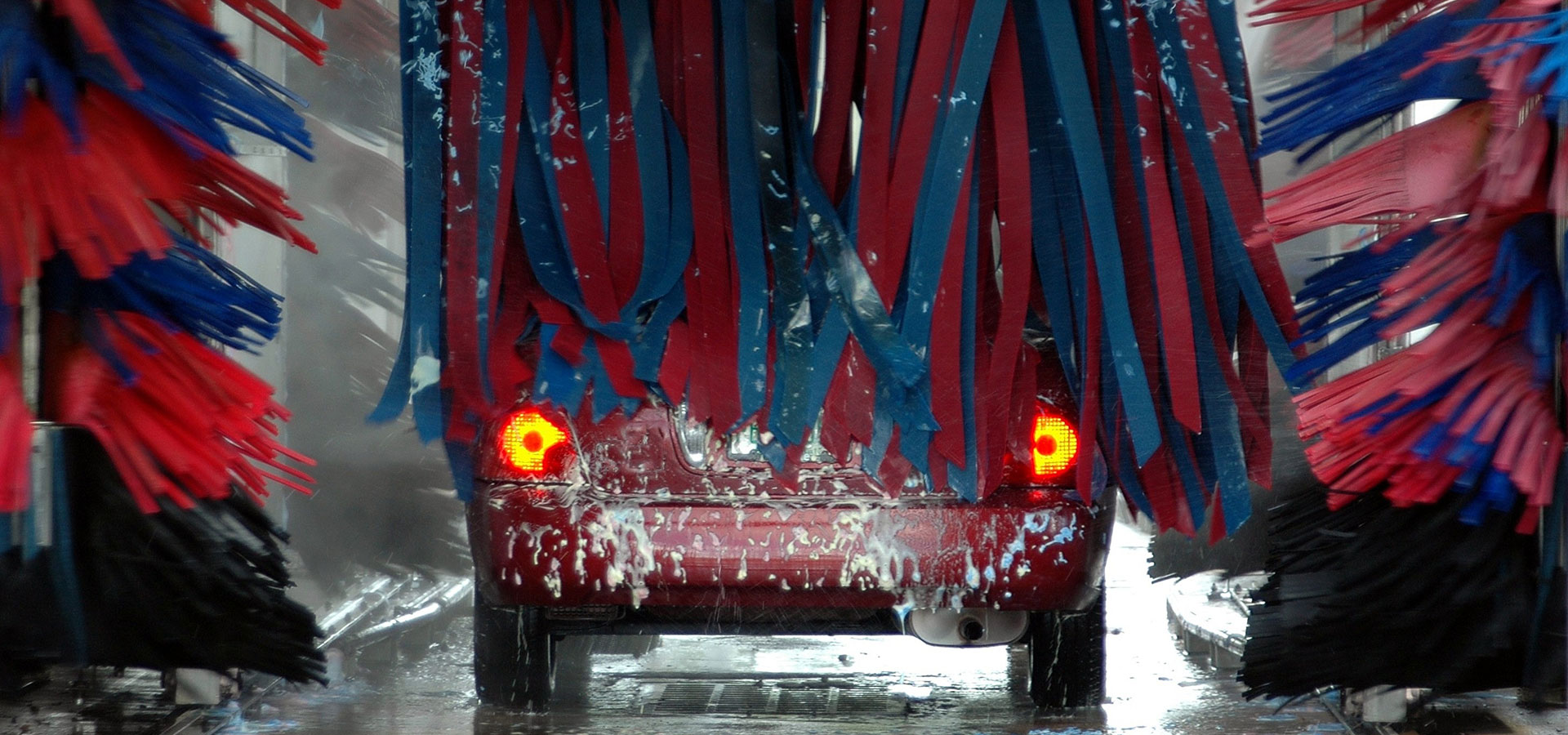 Red car going through an automatic car wash with blue and red cleaning brushes and soap foam